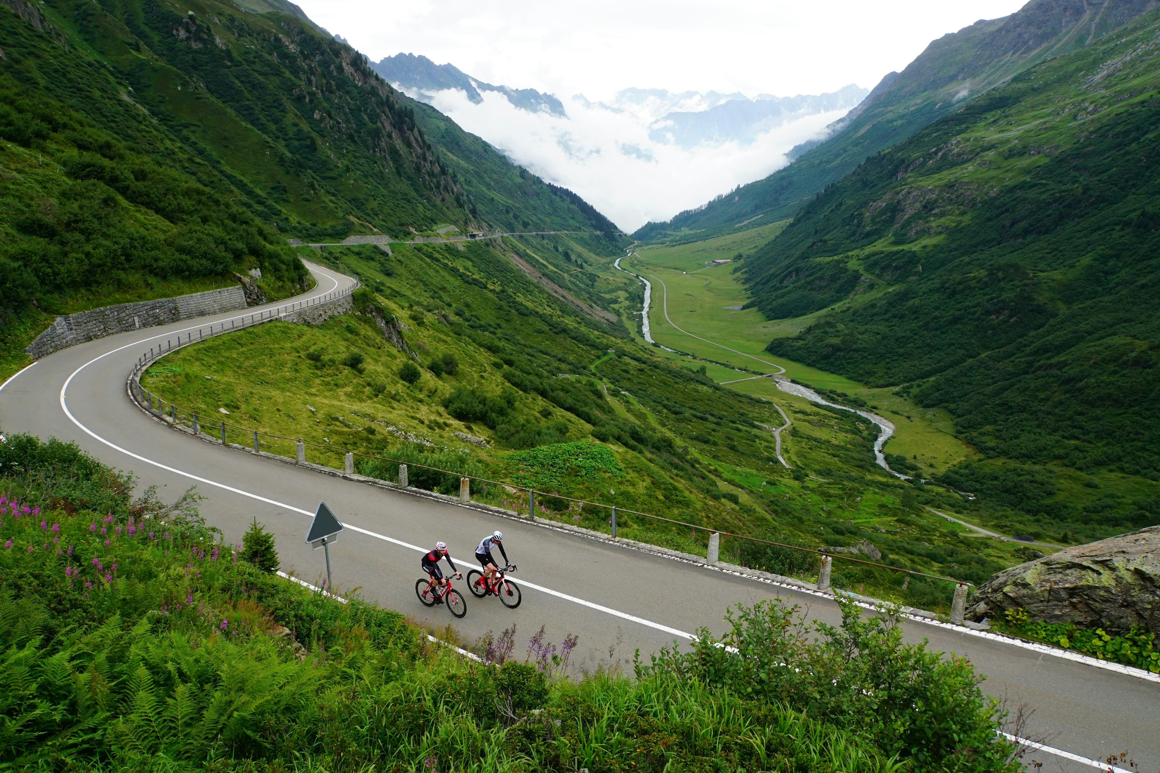 Due ciclisti su una strada di montagna che attraversa una valle verde tra le cime e le nuvole.