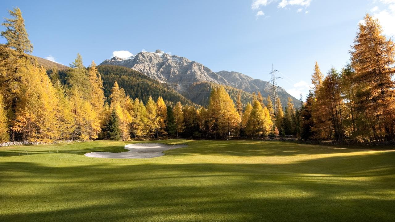 Un campo da golf con bunker di sabbia, circondato da larici dorati e montagne.
