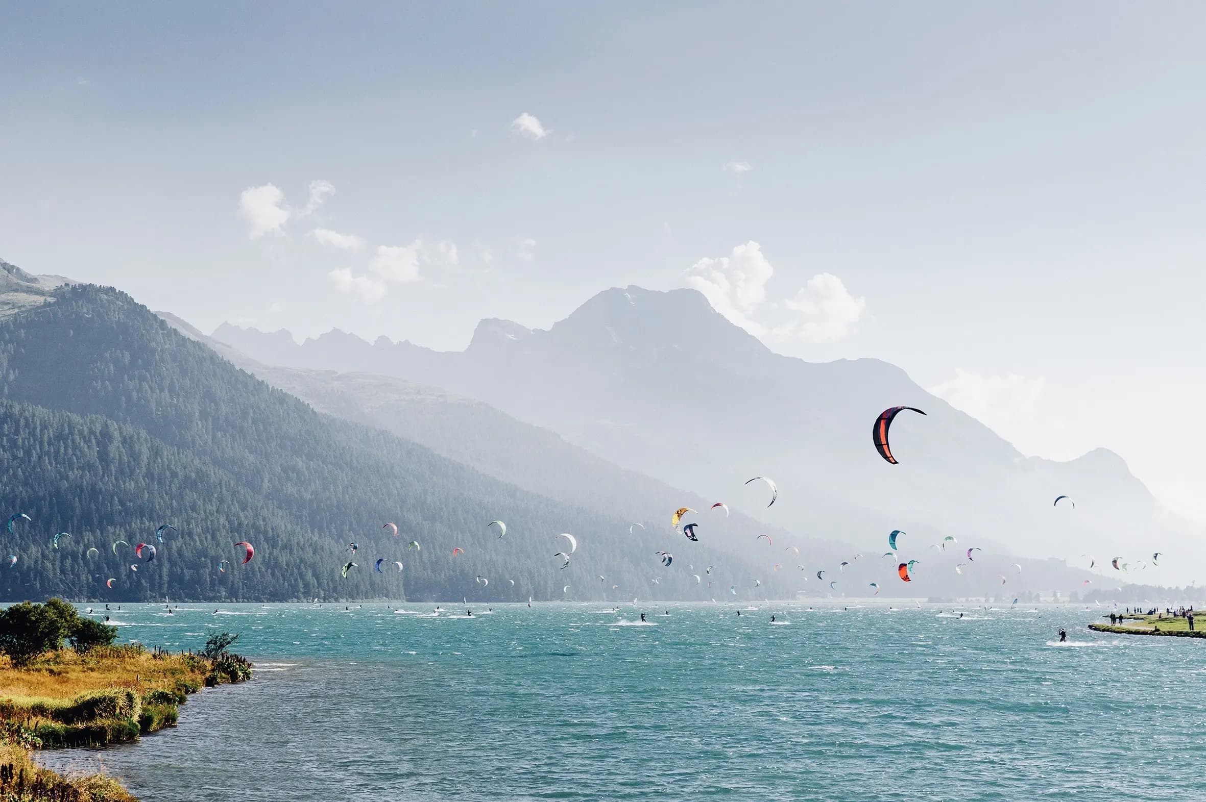 Numerose vele colorate da kitesurf sopra un lago turchese, tra boschi e montagne.