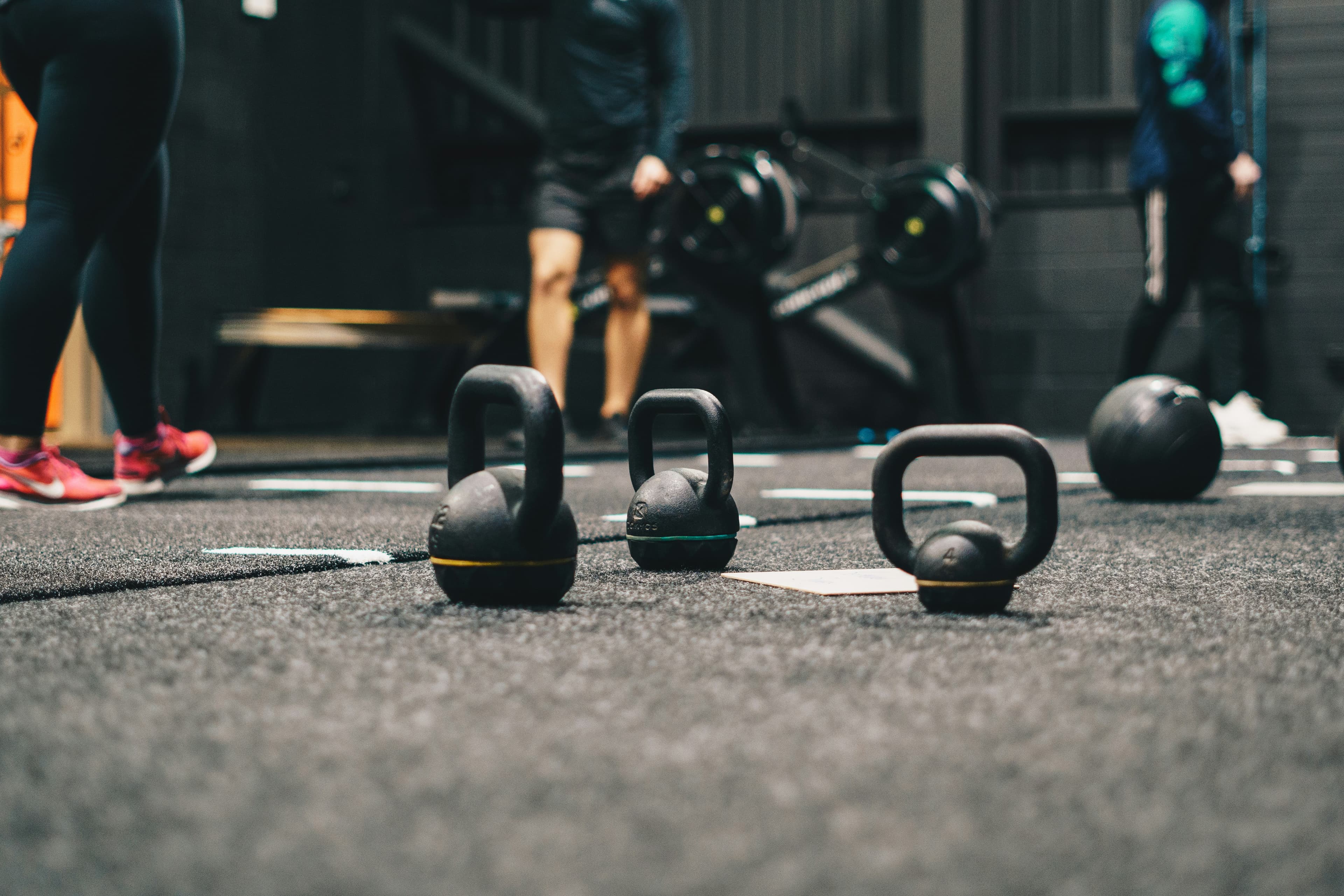 Kettlebell sul pavimento di una palestra, con persone e vogatori sullo sfondo.