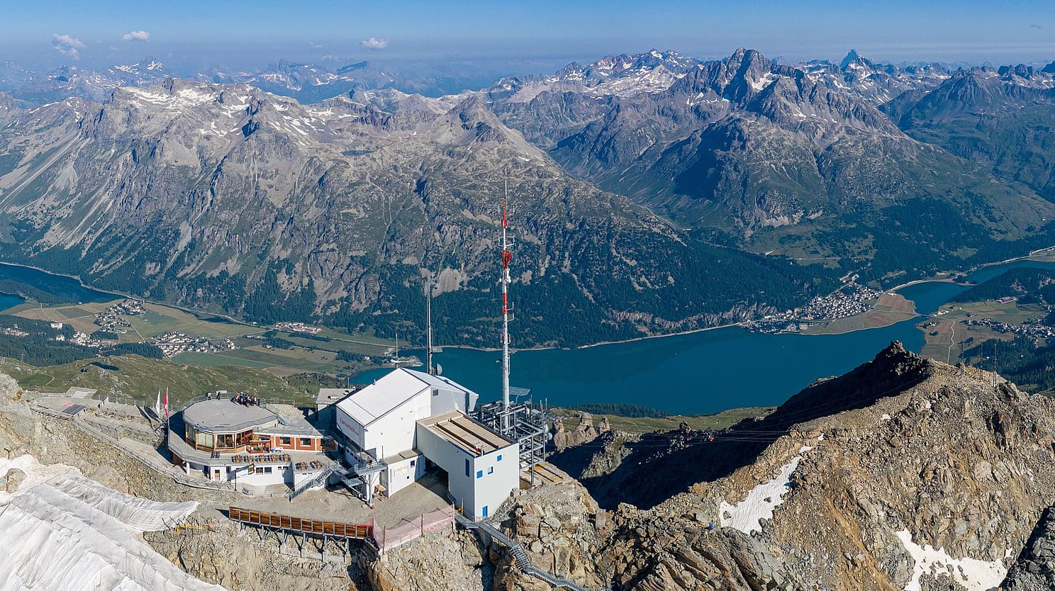 La stazione in quota tra rocce e neve, con il lago e le montagne della valle sullo sfondo.