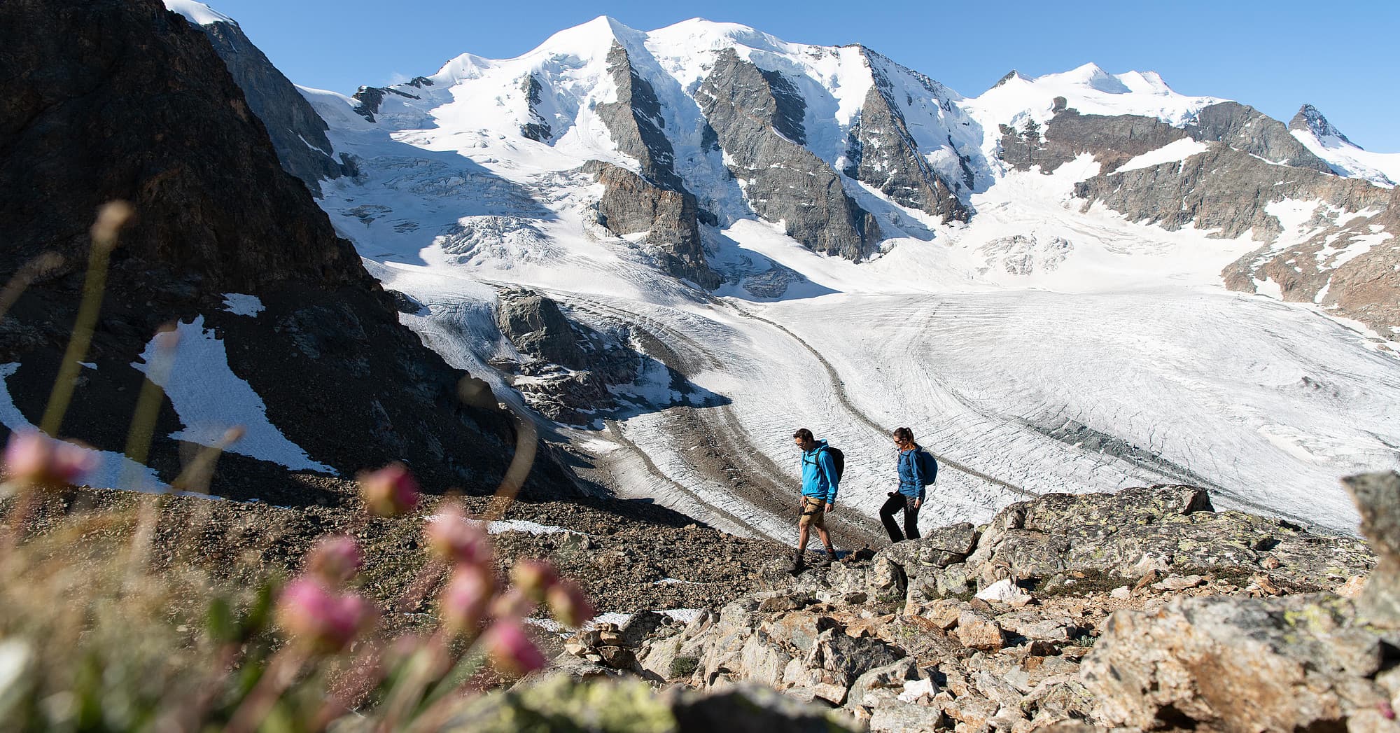 Due escursionisti su un sentiero roccioso, con il ghiacciaio e le cime innevate sullo sfondo.