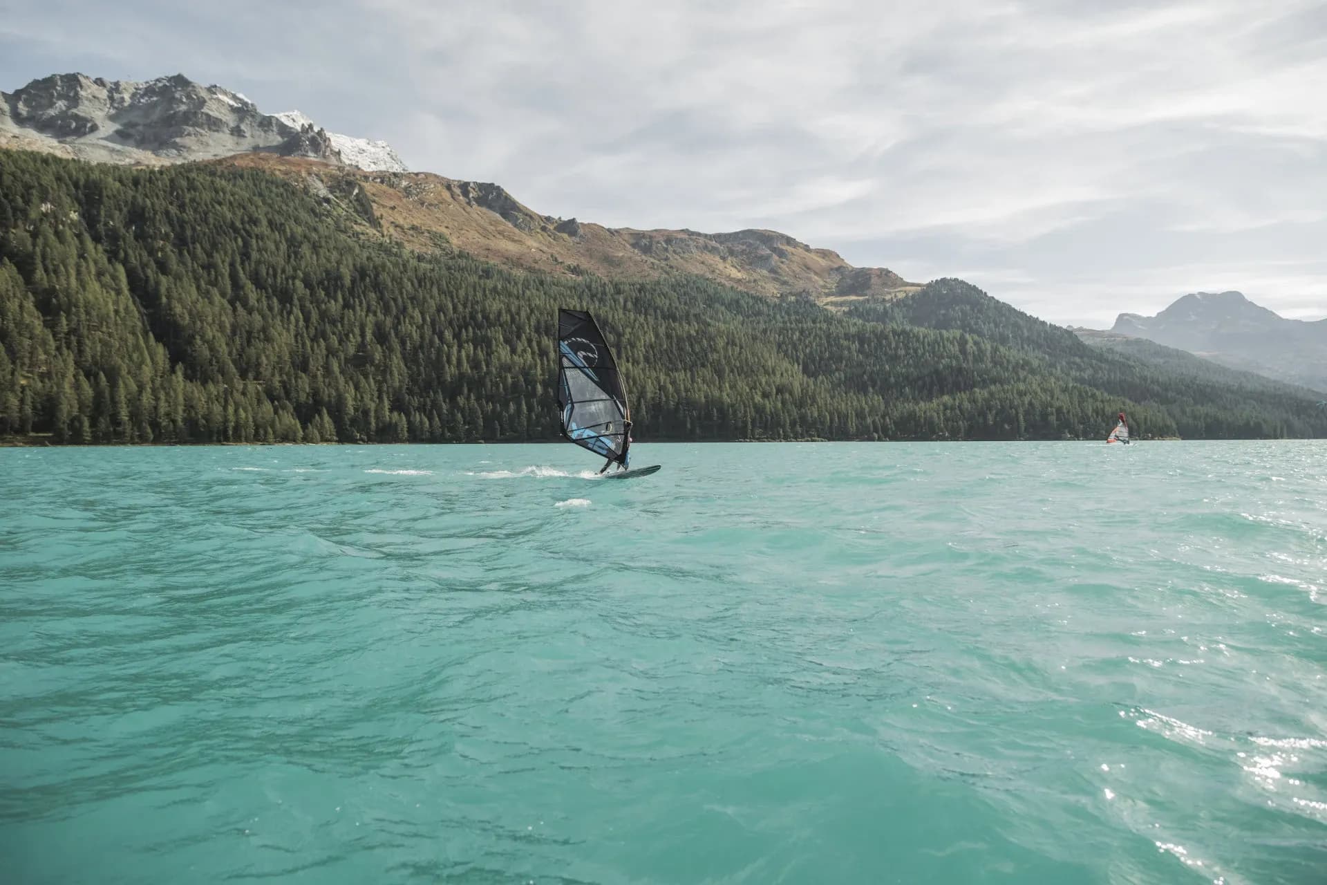 Windsurf sulle acque turchesi di un lago, con boschi e montagne sullo sfondo.